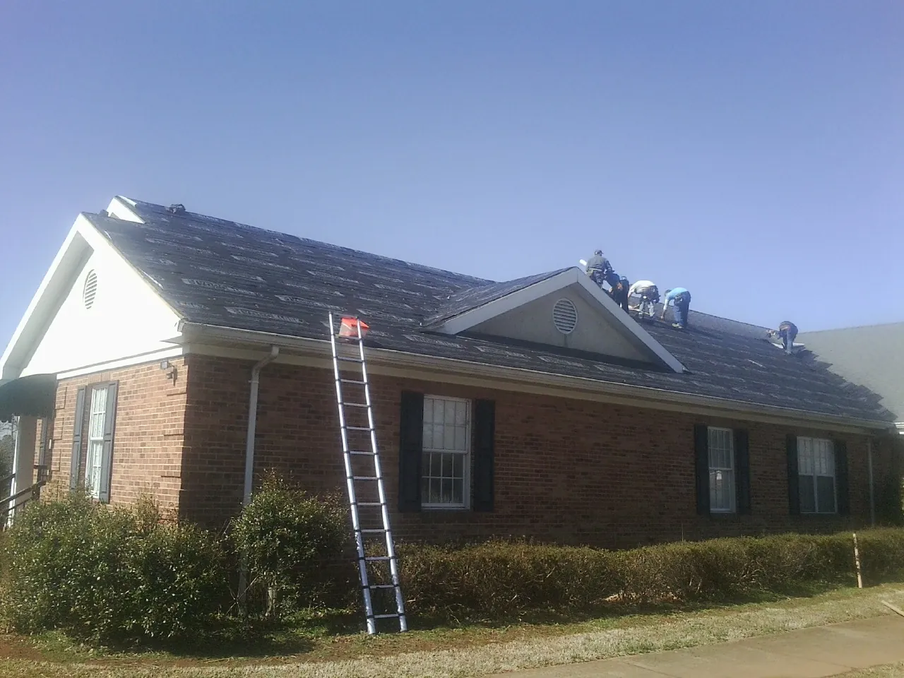 Crew tearing shingles off a brick ranch roof with a ladder set — Emergency Roof Repair work by our crew in Manns Choice
