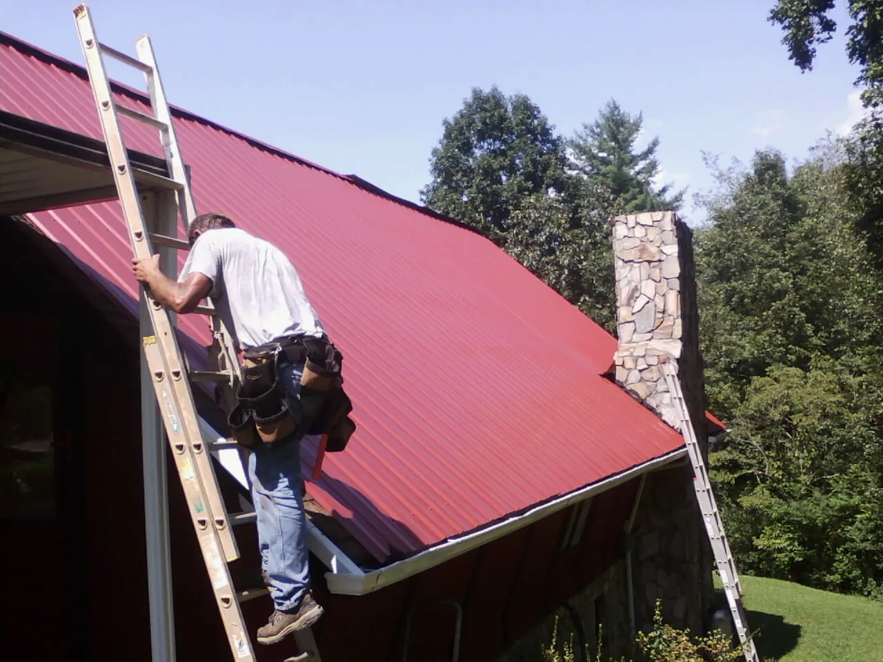 Roofer on a ladder beside a red metal roof and stone chimney — Soffit & Fascia Repair by Roofing Brothers in Manns Choice